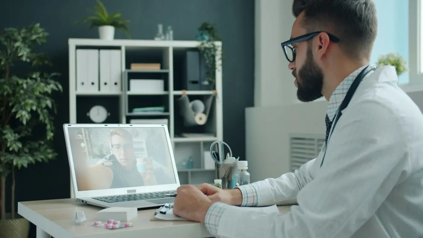 A doctor in a white coat talks with a patient on a laptop video call